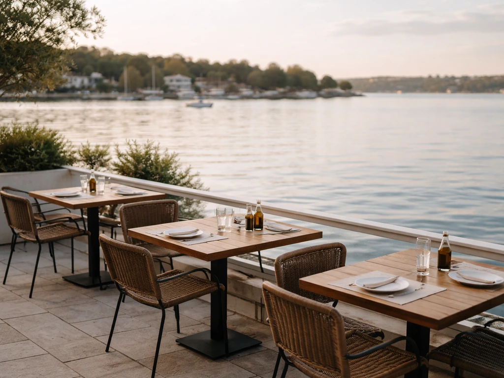 Sunlit waterfront patio with tables and chairs facing calm harbor water and a distant shoreline.