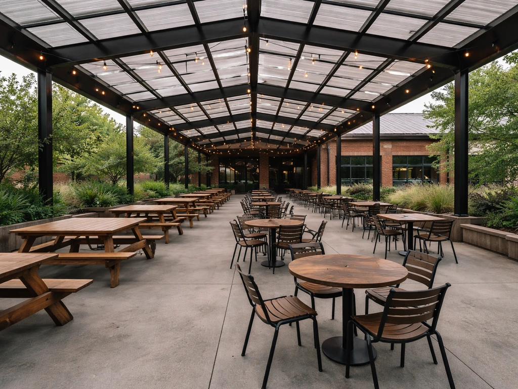 Wide view of a large covered brewery patio with seating, roof, and greenery in natural daylight.